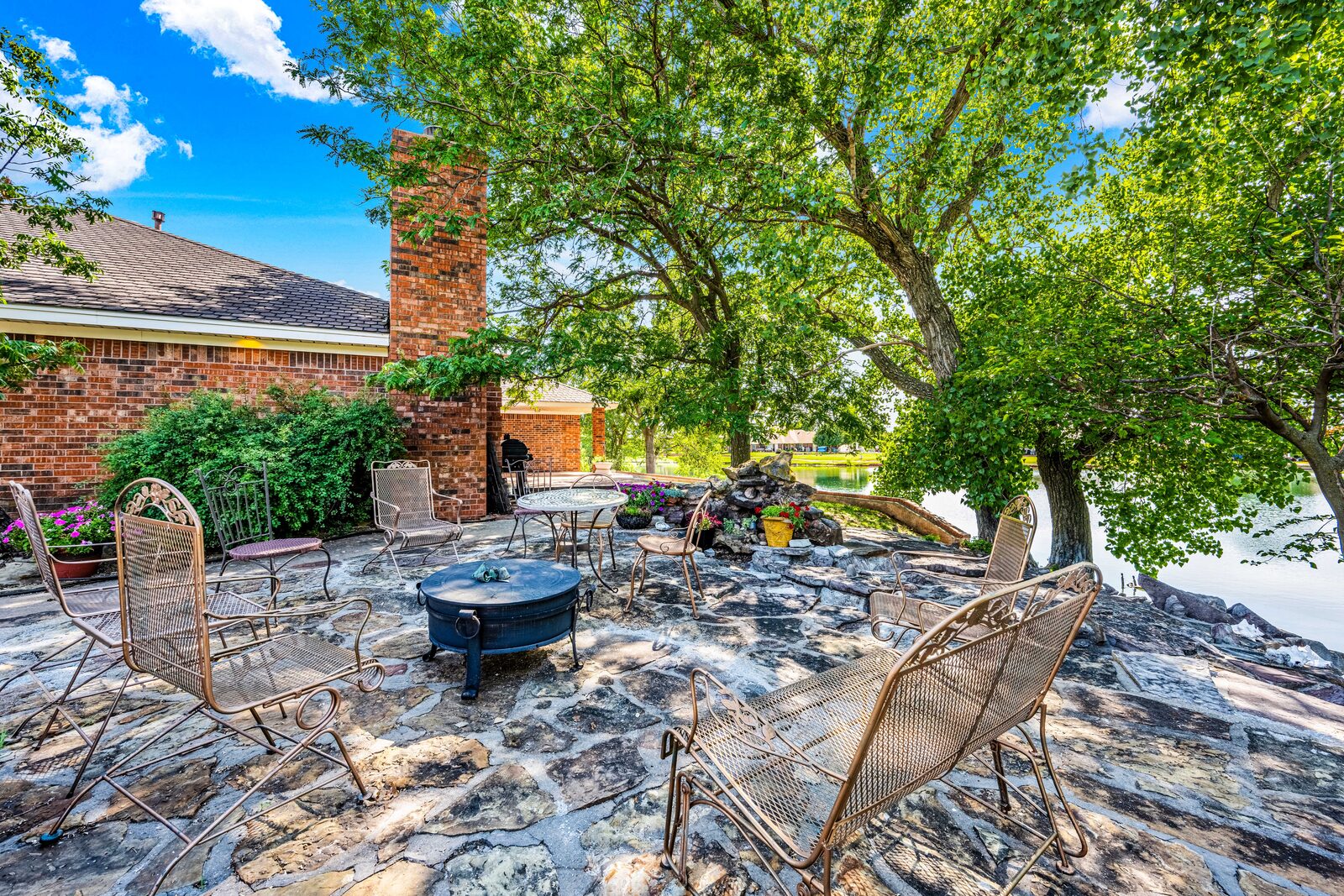 Stone patio at the rear of the home — fire pit, wrought iron furniture, mature trees overlooking the water