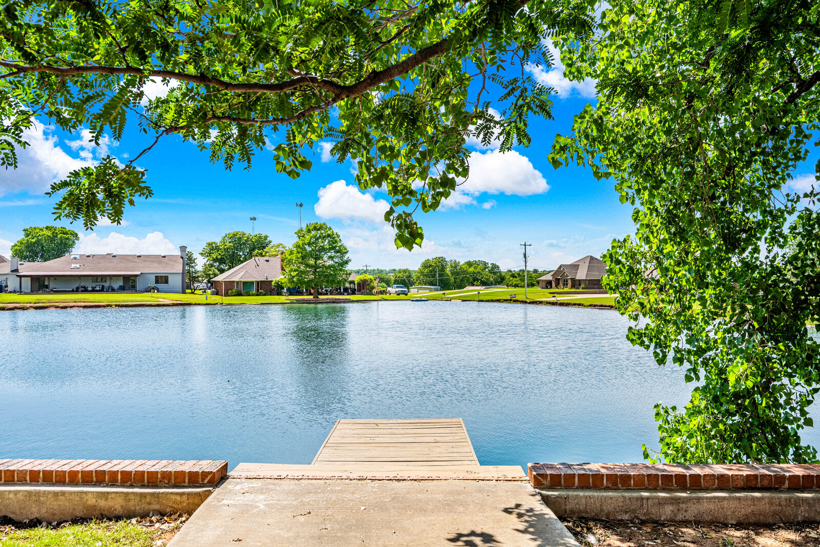 View across the water from the property's dock — mature trees framing the pond with surrounding homes in the distance