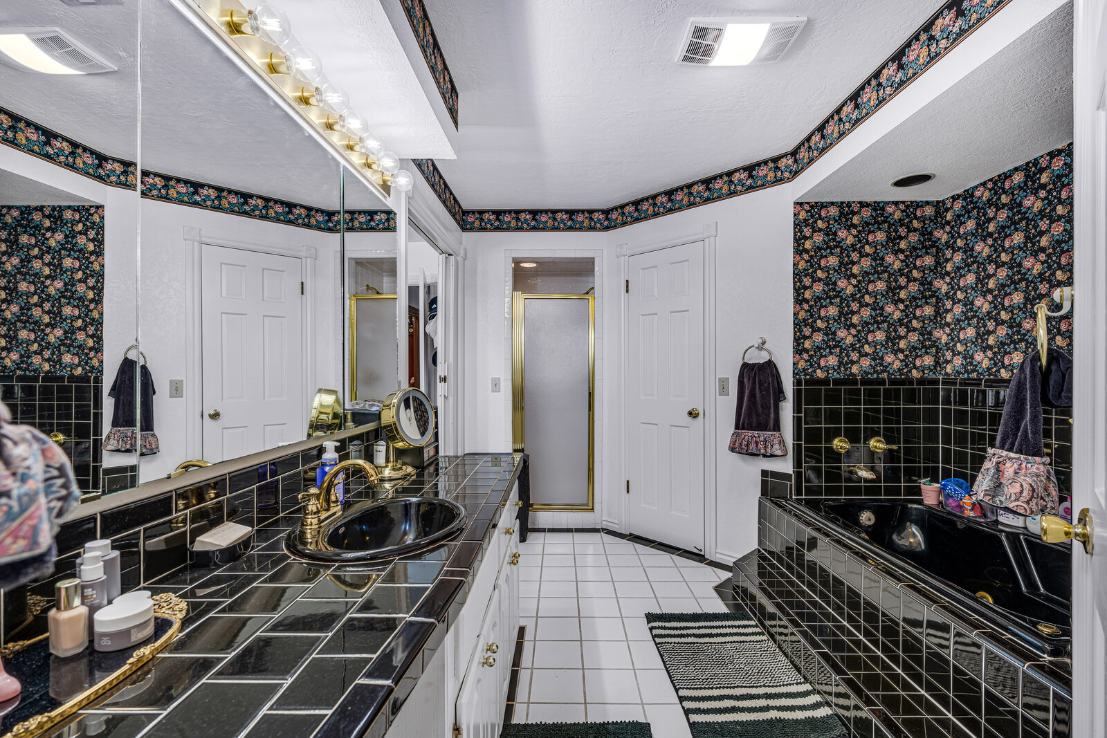 Primary bathroom with black tile countertops and tub surround, brass fixtures, mirrored vanity, and Hollywood-style light bar