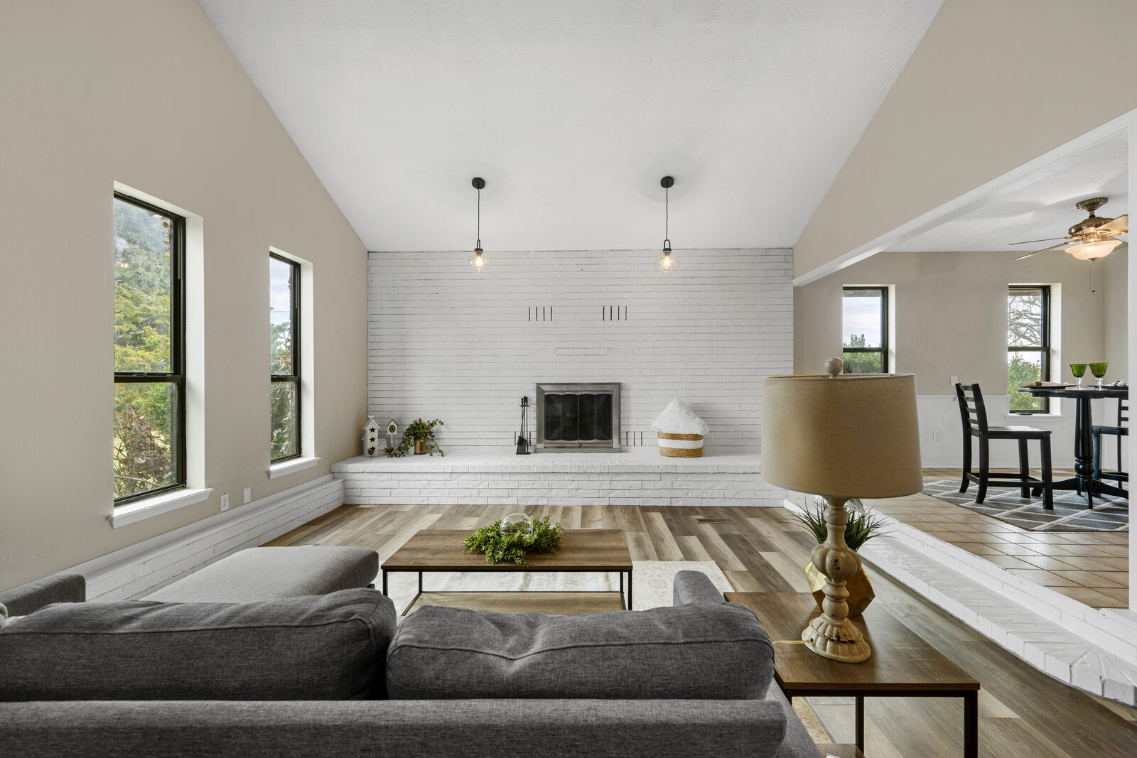 Wide view of the living room showing the full painted white brick wall with the centered fireplace, two industrial pendant lights, large modern windows, and a raised hearth
