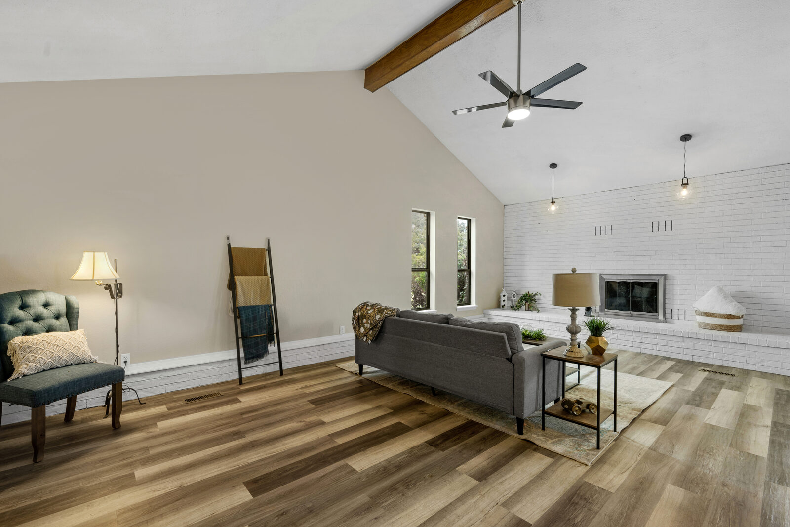 Living room with a vaulted ceiling and a single exposed wood beam — painted white brick fireplace wall, modern industrial pendants, refreshed wood-look flooring