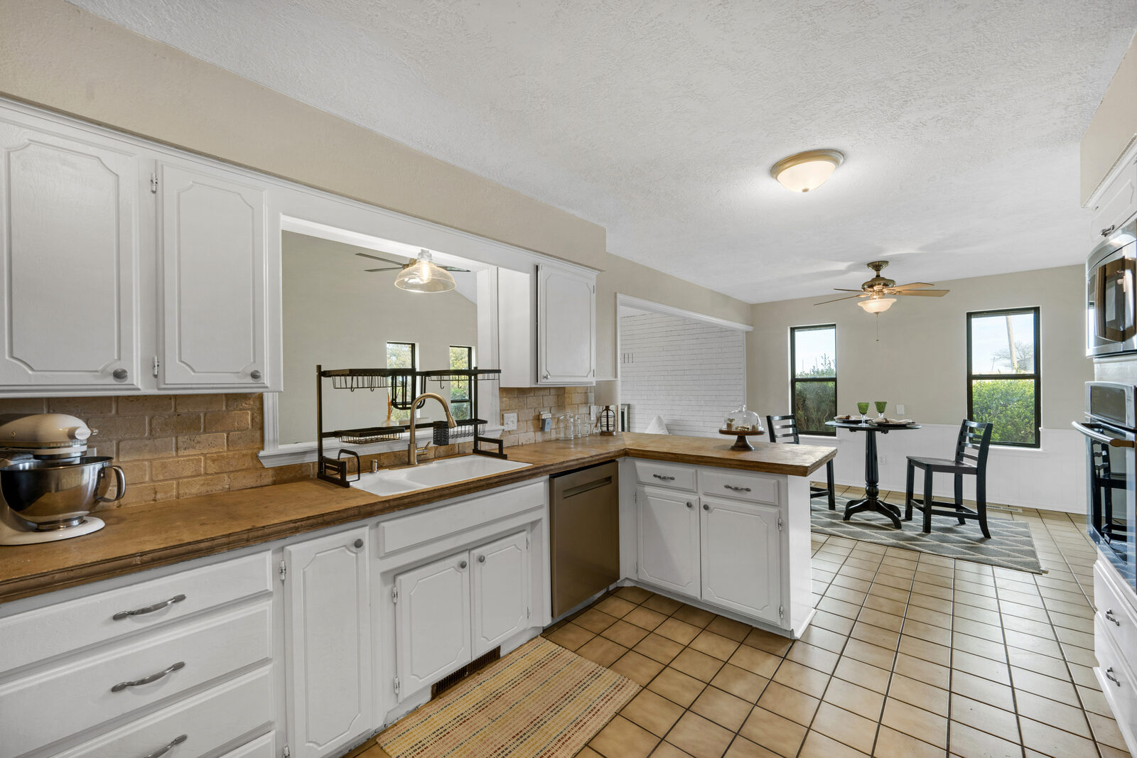 Kitchen with white cabinetry, butcher-block-style counters, brick-style backsplash, stainless dishwasher, and a pass-through window to the living area