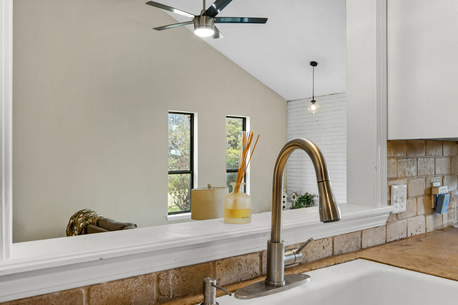 Detail of the kitchen pass-through — brushed steel faucet, brick-style backsplash, and a view of the vaulted living room beyond with the painted white brick wall