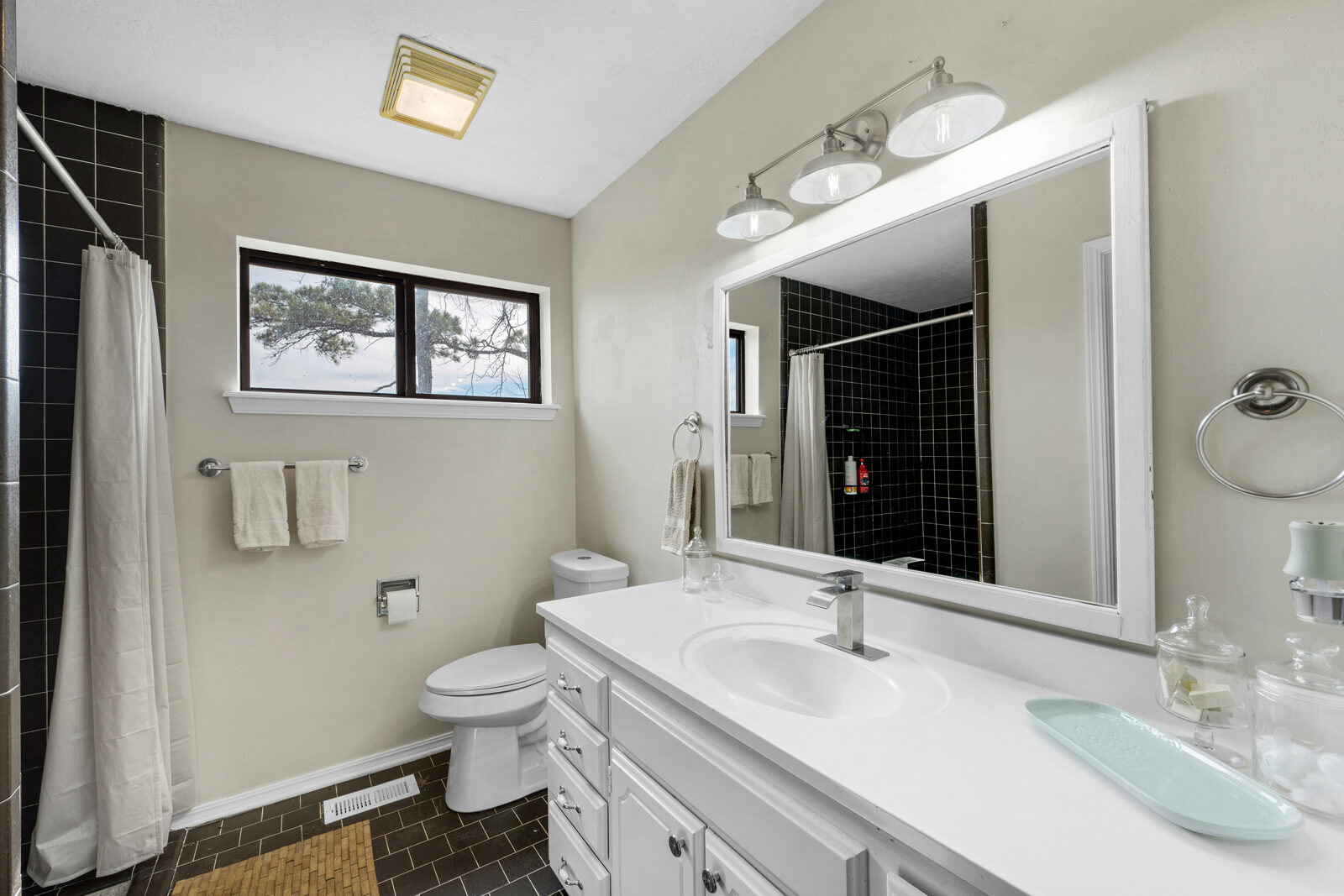 Bathroom with preserved black ceramic tile in the shower and on the floor — modernized vanity with a white wood-framed mirror, three industrial vanity lights, and a window with a rural view