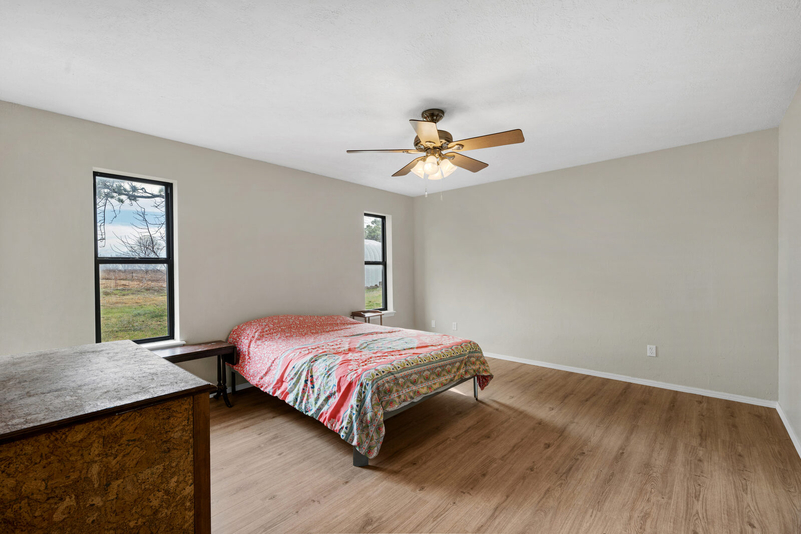 Secondary bedroom with refreshed wood-look flooring, modern black-framed windows looking out to the rural property, and a brass ceiling fan