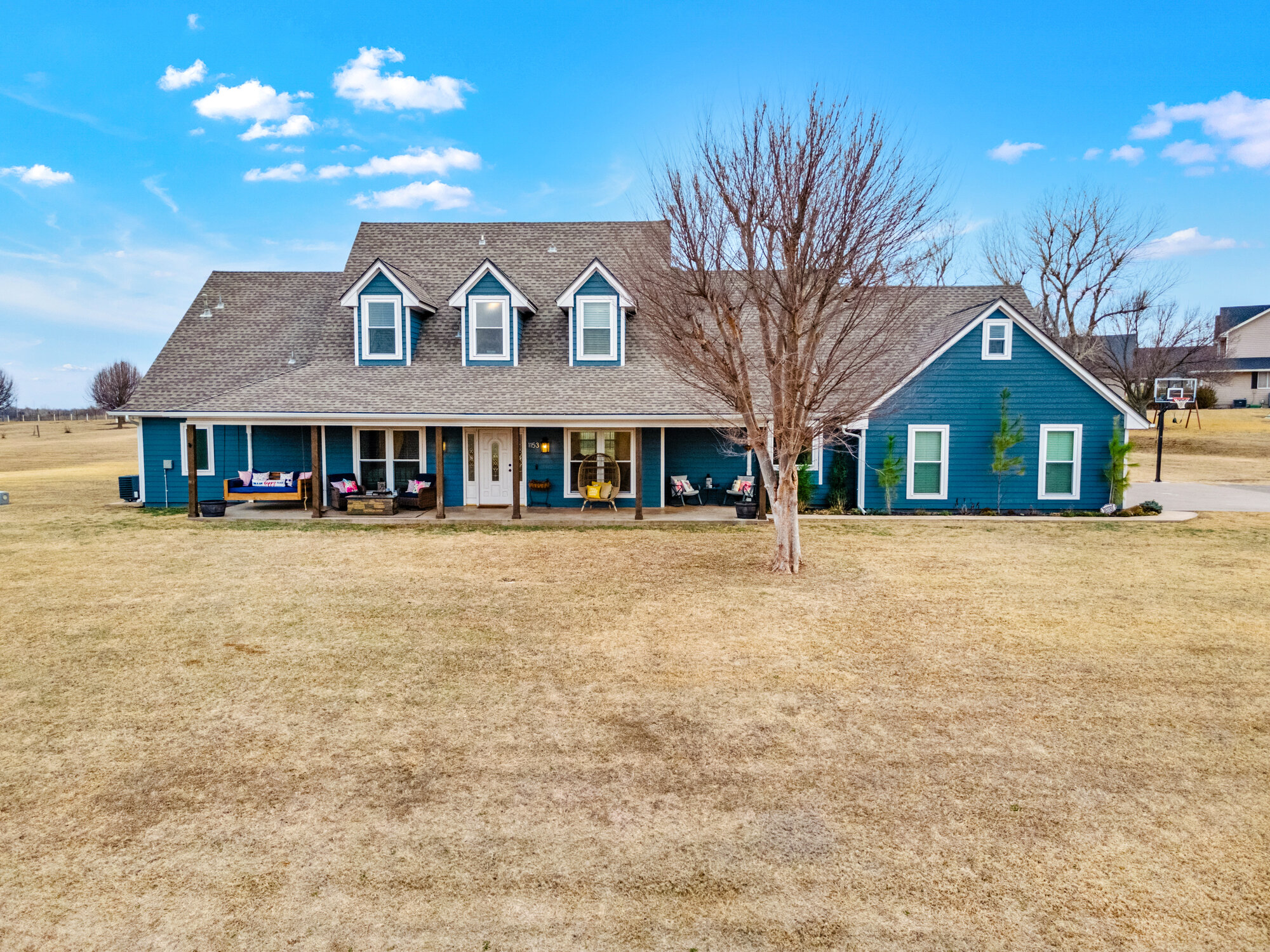 Front exterior of a two-story modern farmhouse in Chickasha — teal-and-white siding, three dormer windows, long covered front porch, captured from a low drone perspective