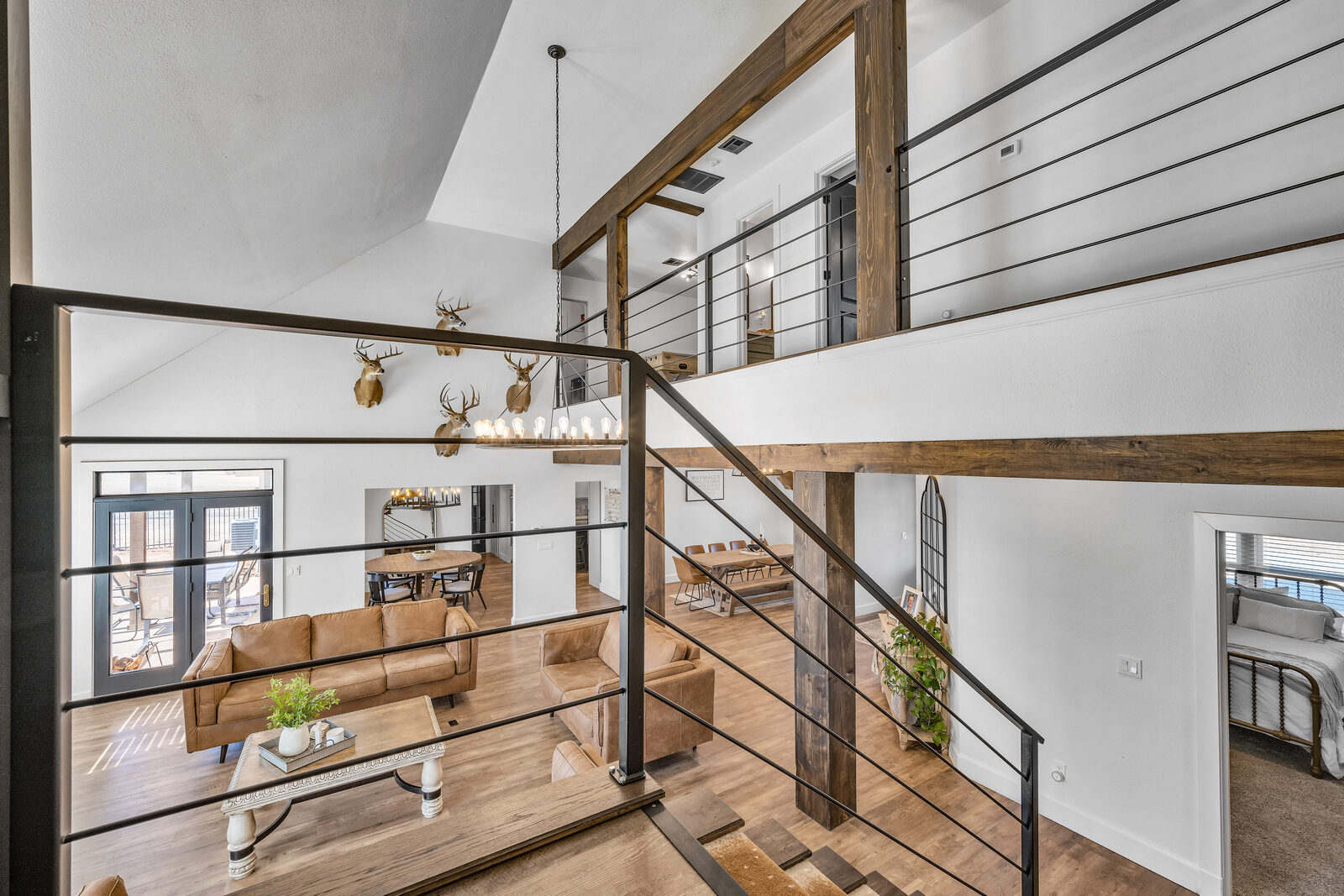 View into the two-story great room from the upper balcony — reclaimed wood beams, modern black metal railings, tan leather seating, dramatic chandelier