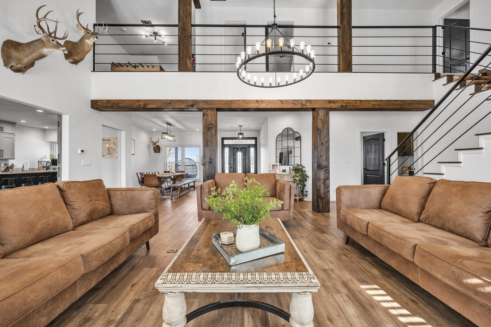 Two-story great room from the main floor — vaulted ceiling, circular Edison-bulb chandelier, reclaimed wood support beams, three tan leather sofas around an antique-style coffee table