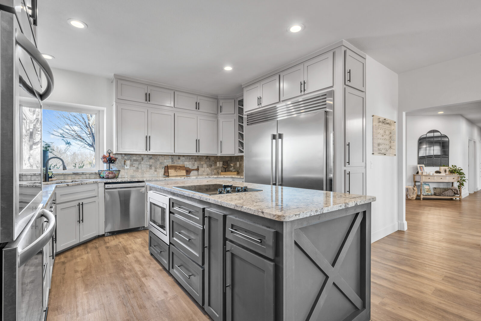 Kitchen with two-tone cabinetry — white perimeter cabinets with a darker gray island, granite counters, travertine backsplash, and built-in stainless refrigerator