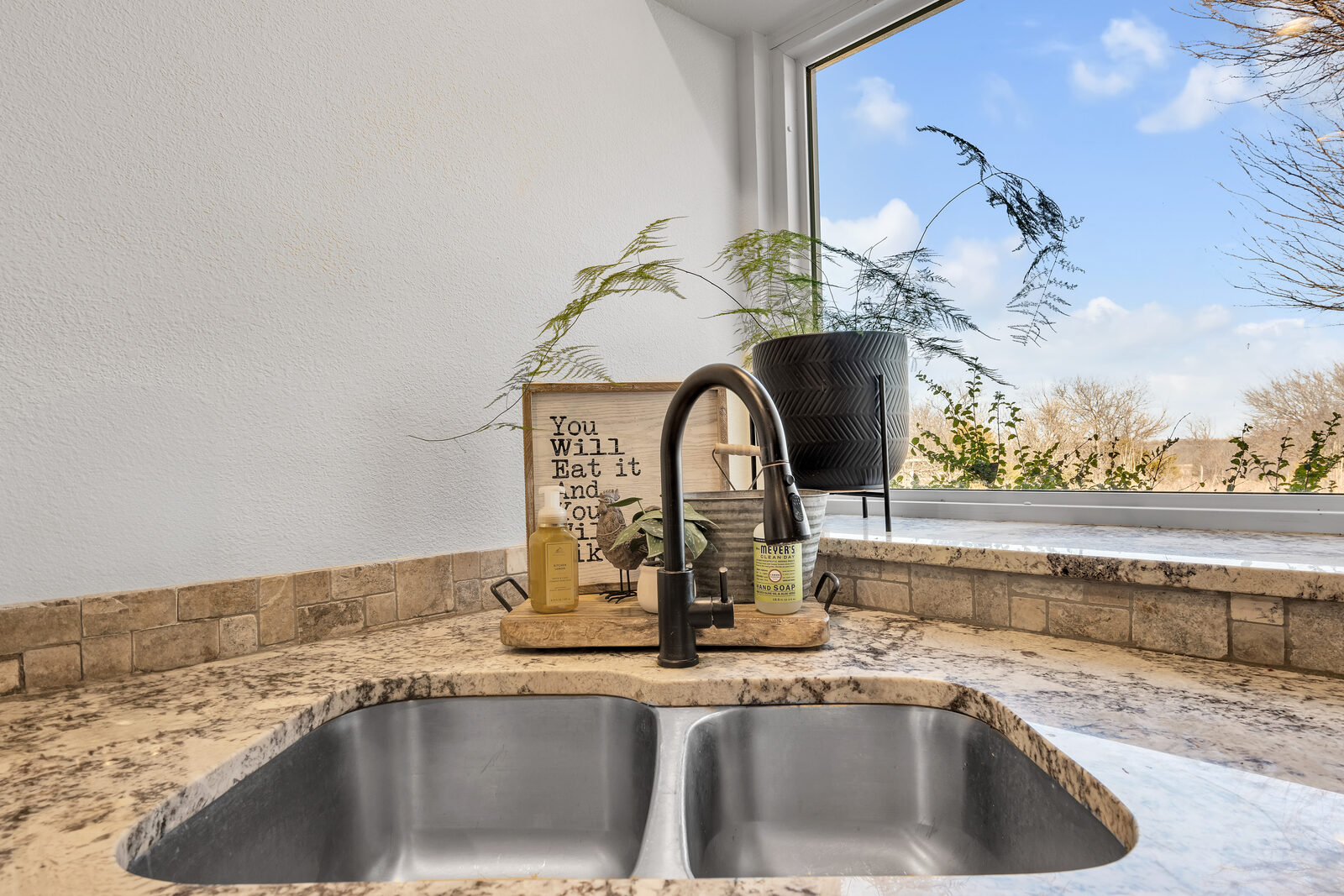 Kitchen sink detail — double-basin undermount sink, oil-rubbed bronze faucet, granite counter, travertine backsplash, large window with a countryside view