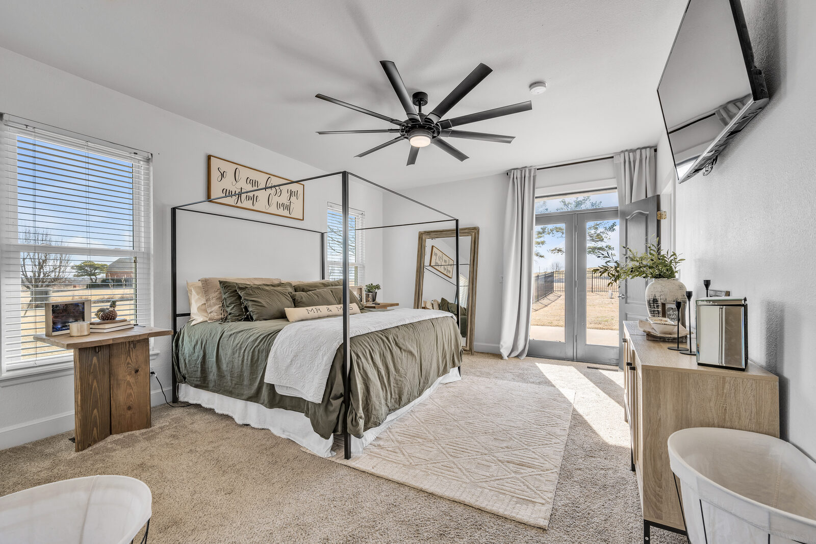Primary bedroom — iron canopy bed, sage and white linens, modern ceiling fan, French doors leading to the backyard with a countryside view