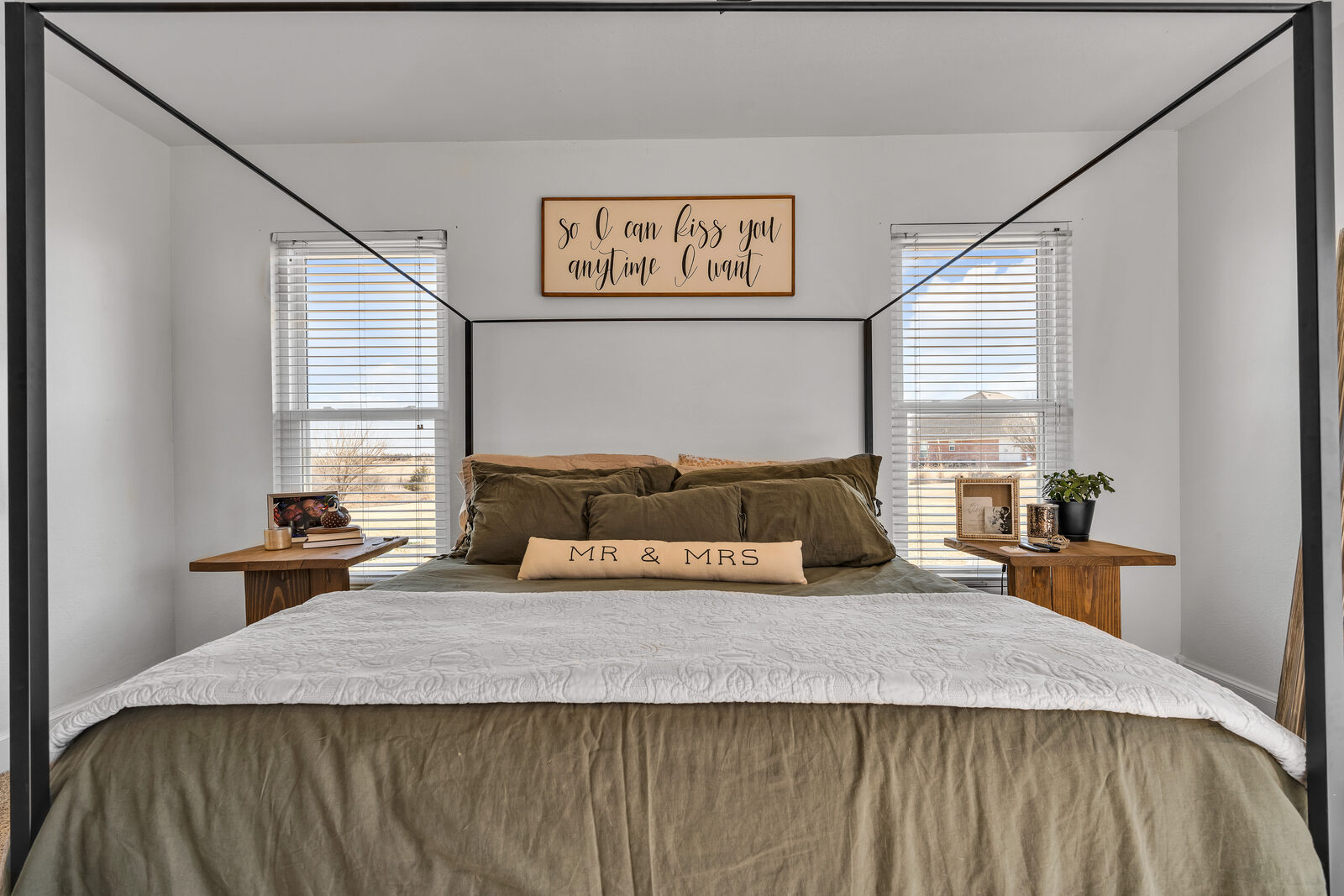 Primary bedroom centered on the bed — iron canopy frame, sage green and white bedding, paired wood nightstands, windows on both sides framing a country view