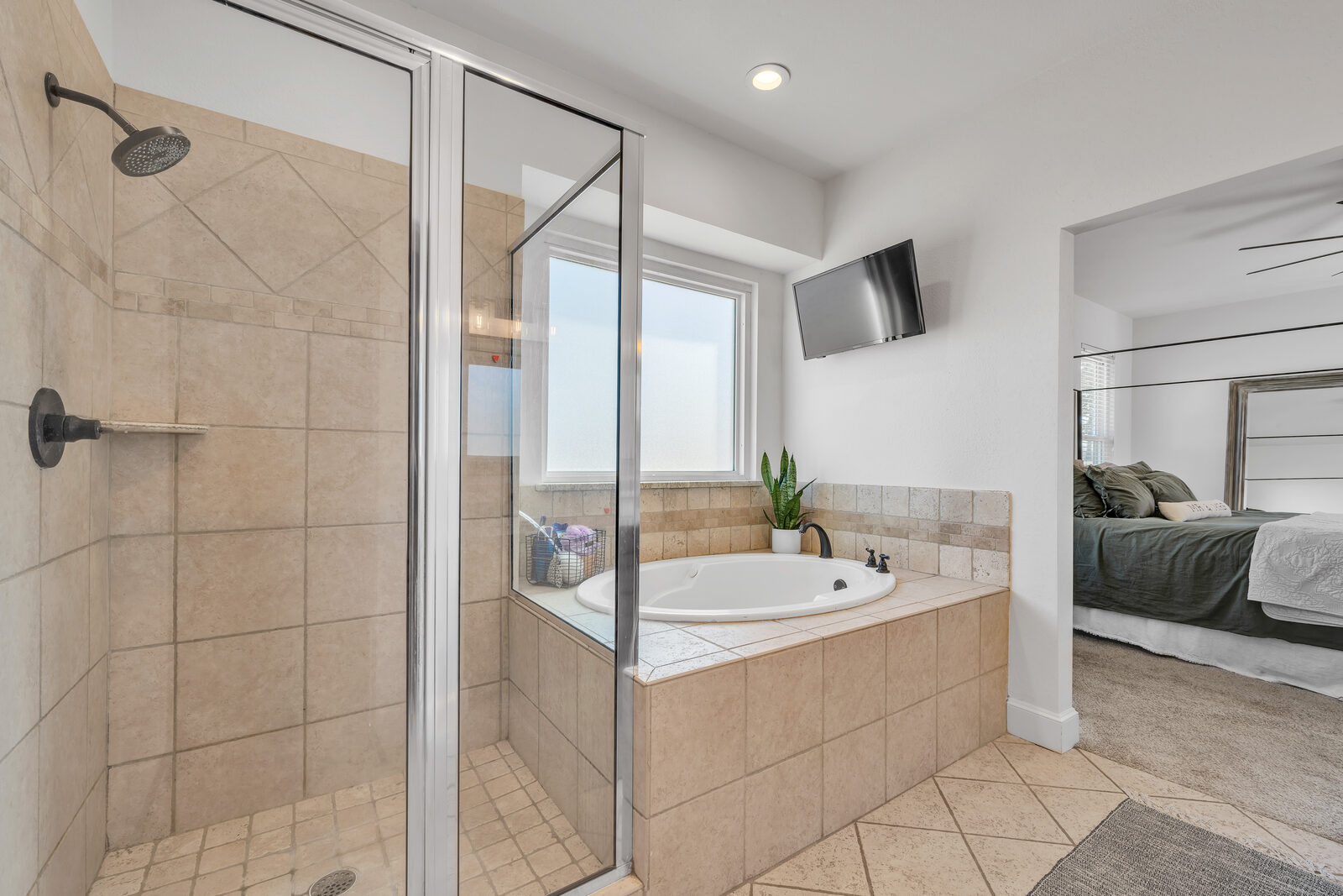 Primary bathroom — walk-in shower with travertine tile and glass enclosure, garden tub with tile surround, oil-rubbed bronze fixtures, view through to the bedroom
