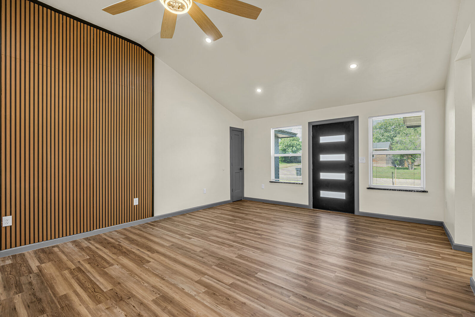 Living room looking toward the front entry — modern dark glass door, wood slat accent wall, vaulted ceiling, light-toned wood flooring