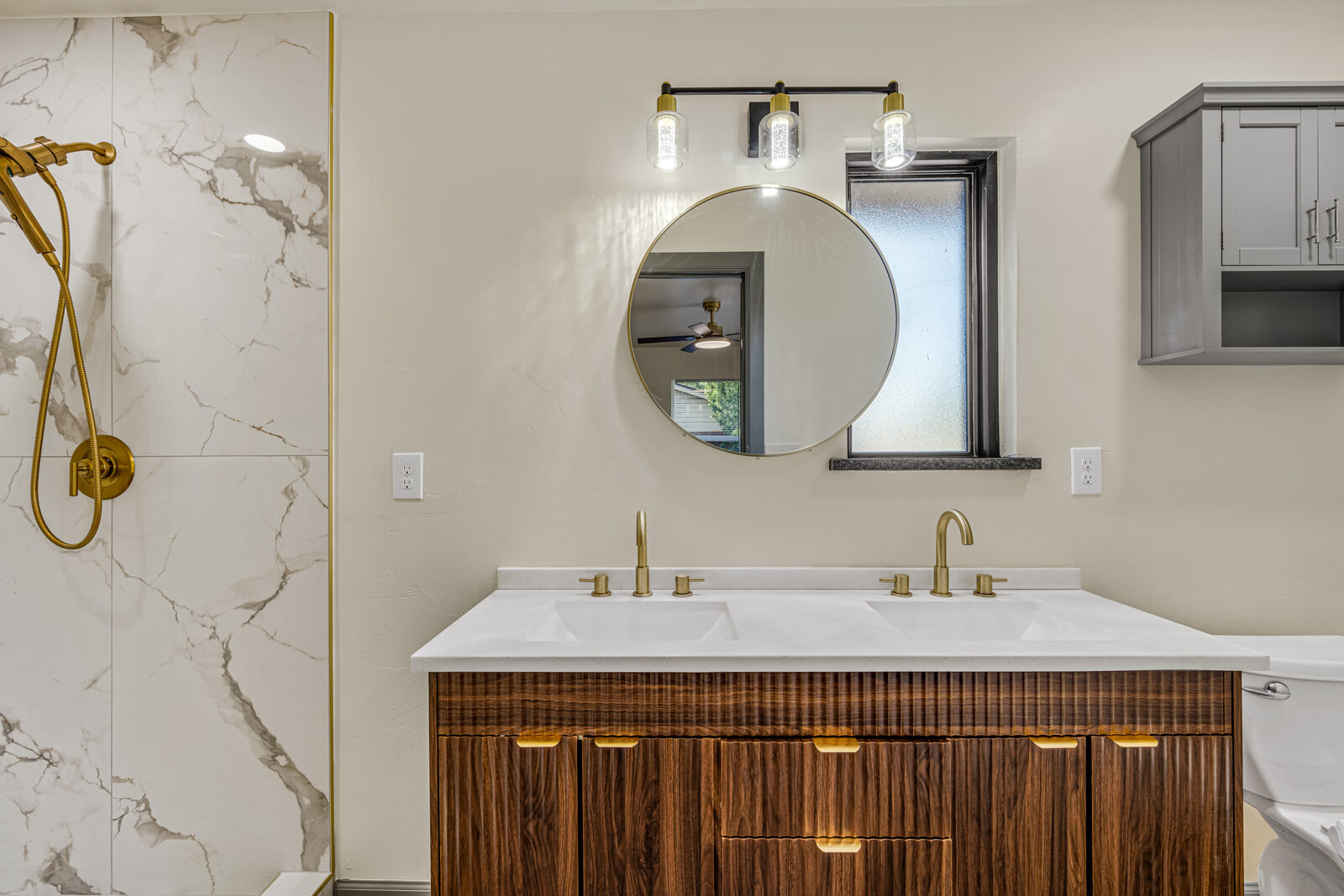Primary bathroom — marble shower surround with brass fixtures, walnut vanity with fluted fronts, round mirror, quartz countertop