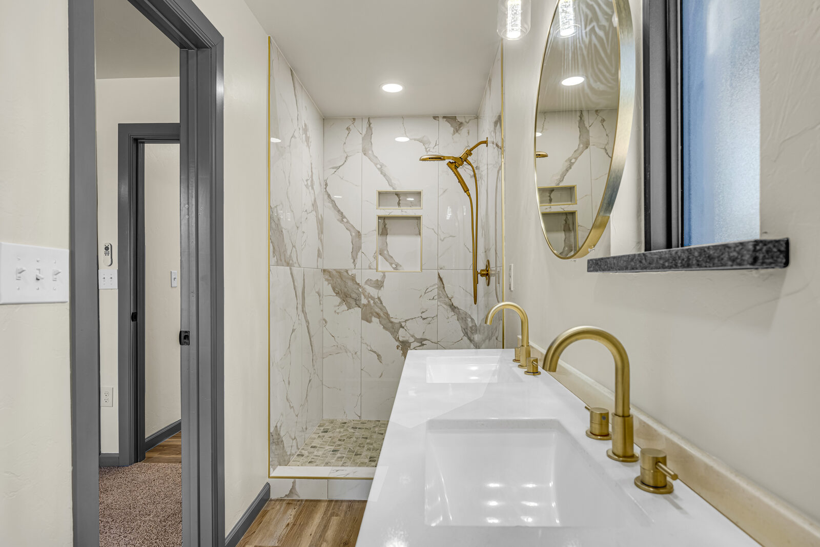 Primary bathroom vanity detail — long quartz top with dual sinks, brass faucets and shower hardware against the marble surround