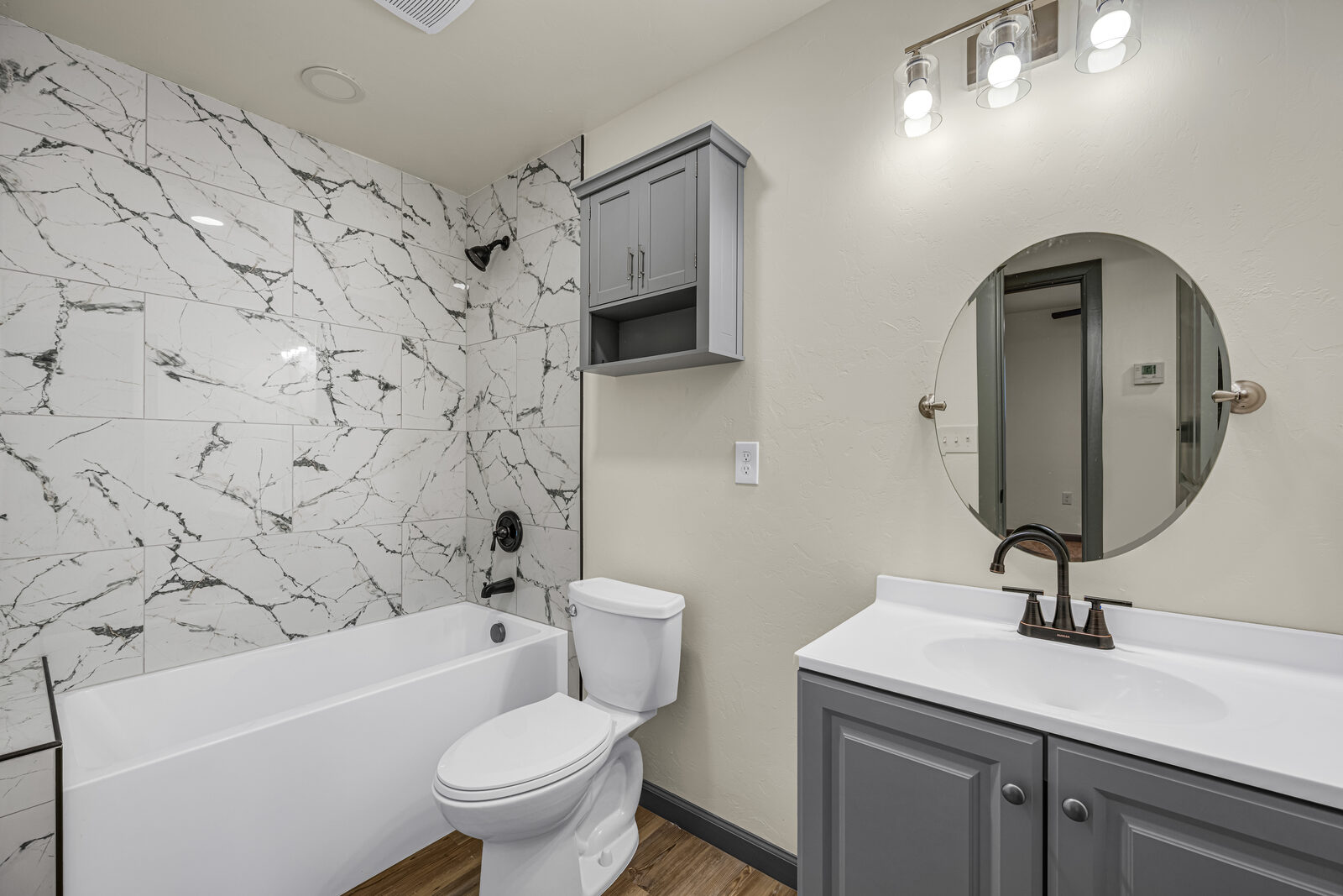 Secondary bathroom — marble tile shower surround, gray vanity, white quartz countertop, round mirror, satin bronze faucet