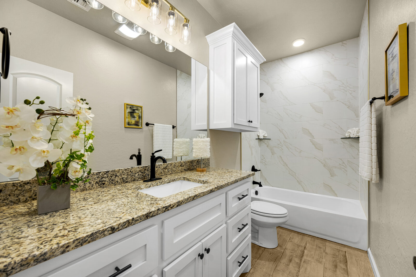 Secondary bathroom with white cabinetry, granite vanity, marble-look tile tub surround, and wood-look tile flooring