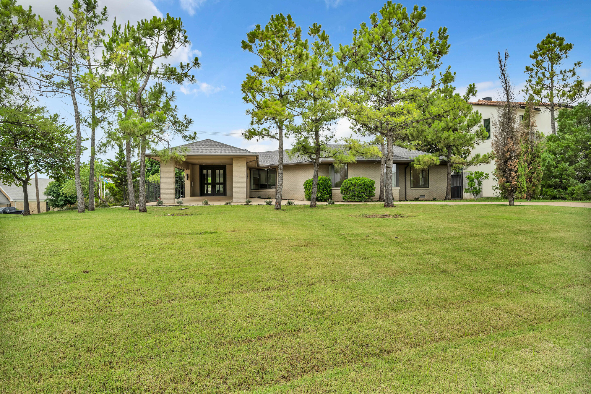 Front exterior of a renovated single-story modern estate in Nichols Hills — cream painted brick, covered entry portico with glass doors, mature pines framing a deep front lawn