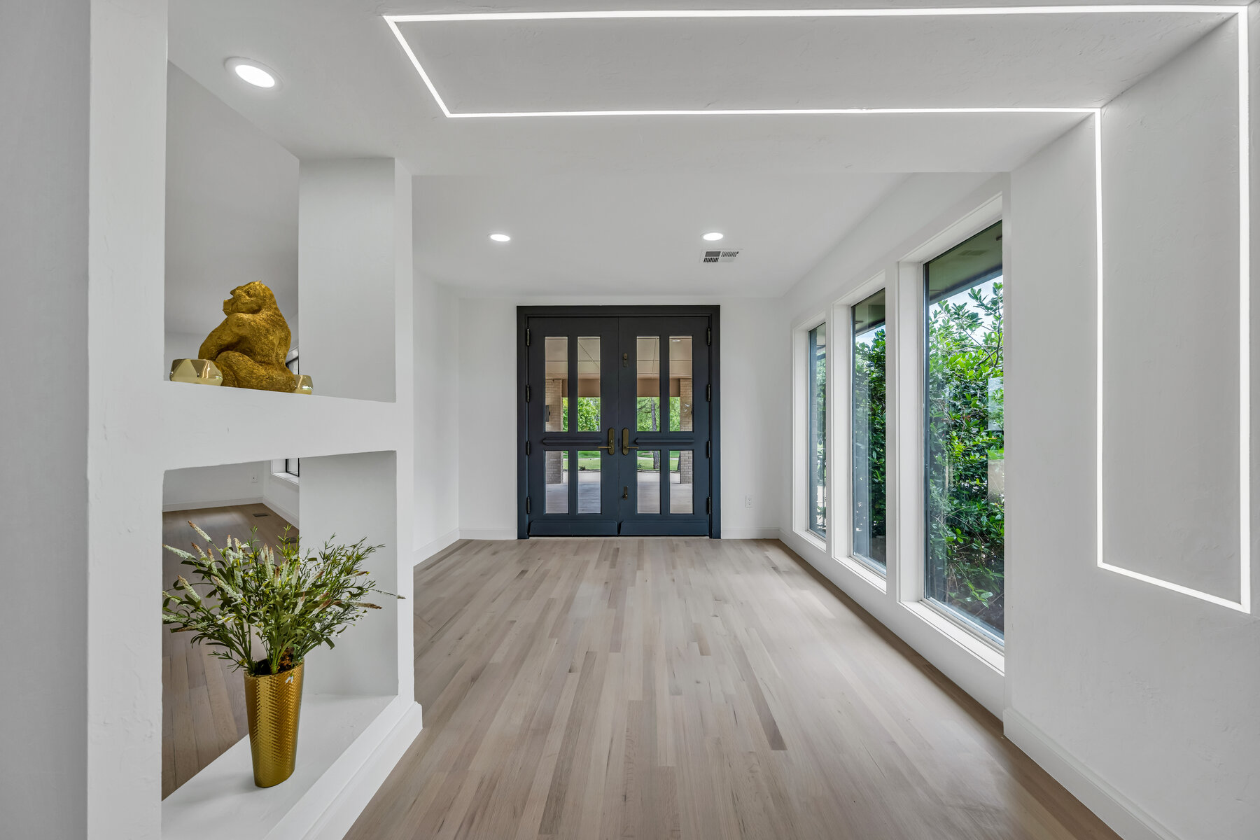 Entry foyer with integrated LED architectural lighting tracing the ceiling and walls — light oak floors, dark double front doors, floor-to-ceiling windows on the right