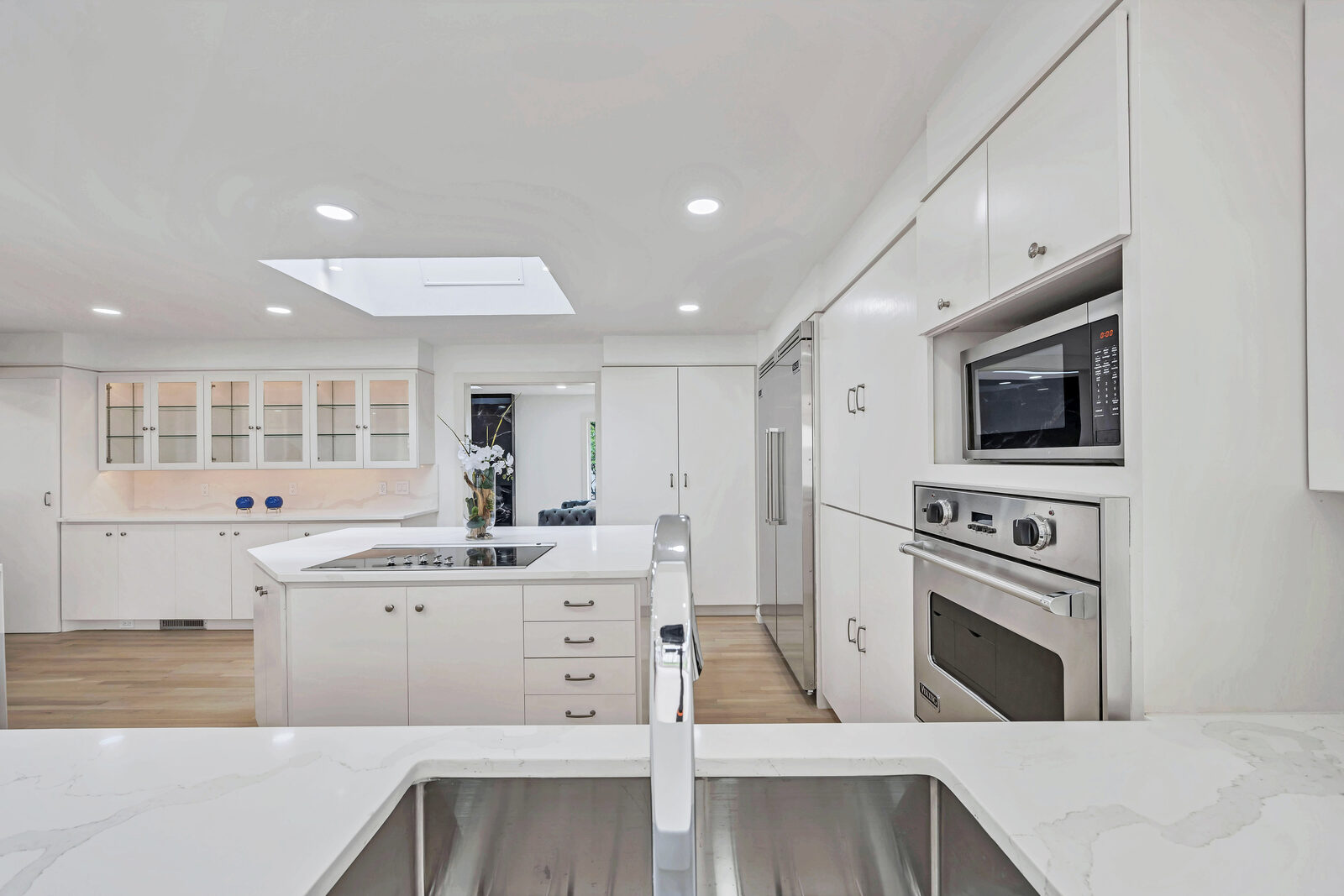 Chef's kitchen with white cabinetry, light maple floors, illuminated glass-front display cabinets, professional built-in oven and microwave, and a center island with cooktop