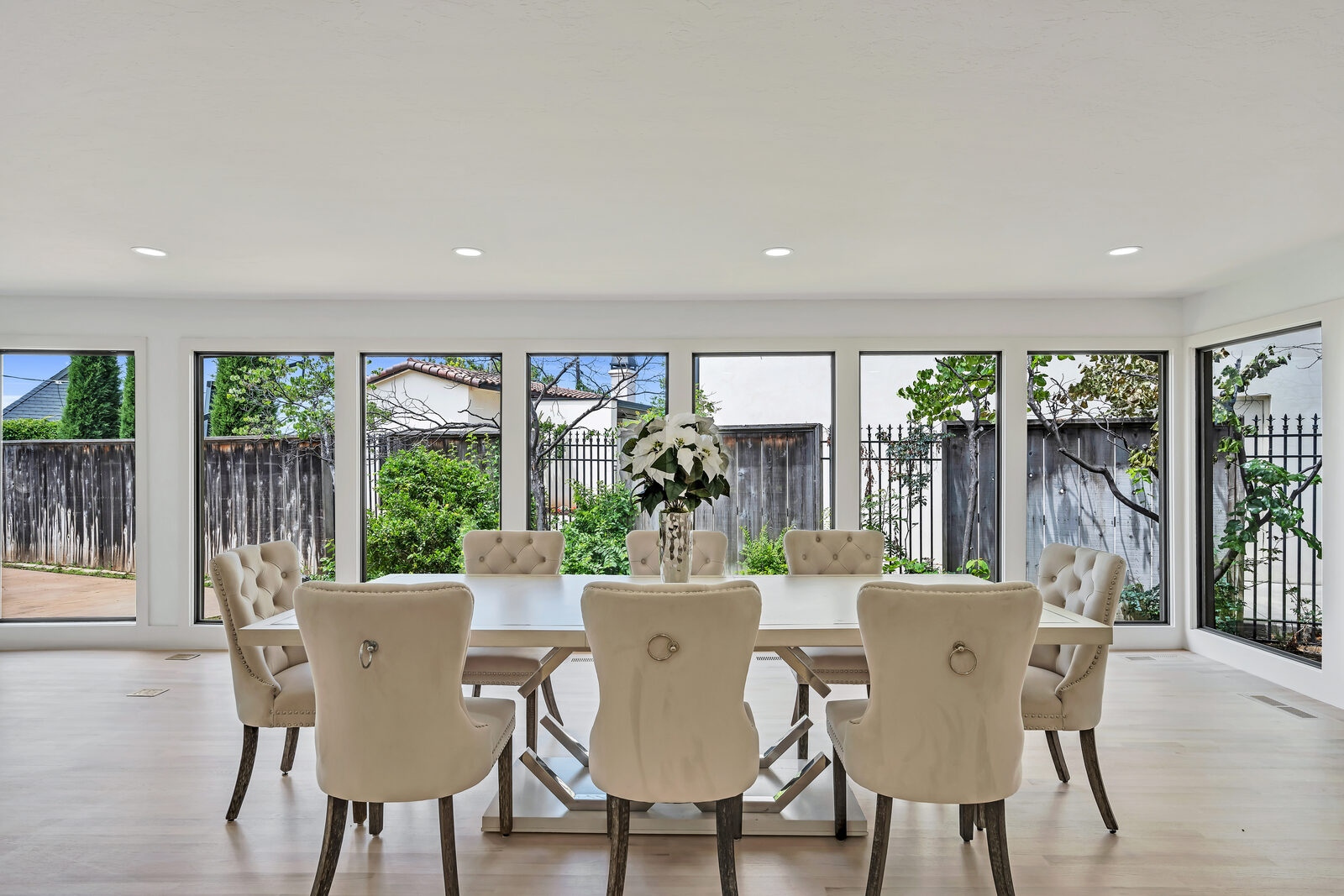 Formal dining room — wraparound wall of black-framed windows looking out onto the gardens, long modern table with eight tufted velvet chairs