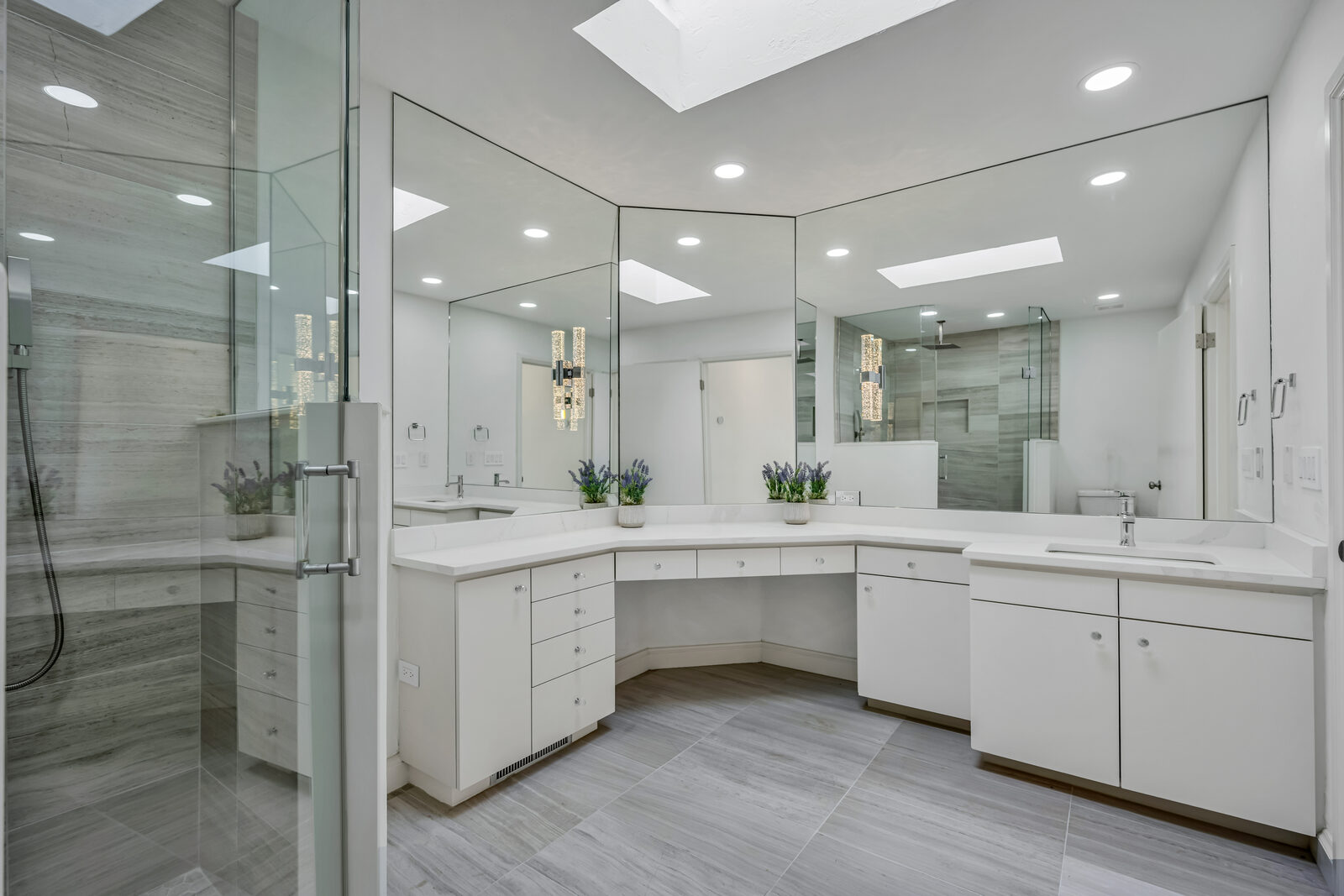 Primary bath with wraparound floor-to-ceiling mirrors, dual skylights, a center makeup vanity flanked by twin sinks, crystal bubble sconces, and a glass-enclosed shower with gray stone-look tile