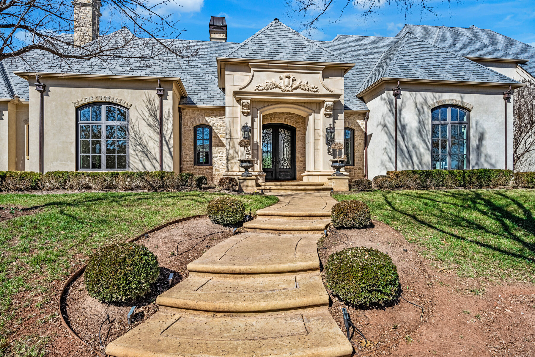 Front entry detail — carved limestone portal with ornate cartouche, wrought iron and glass double doors, stone columns, arched windows flanking the entry, manicured boxwood landscaping