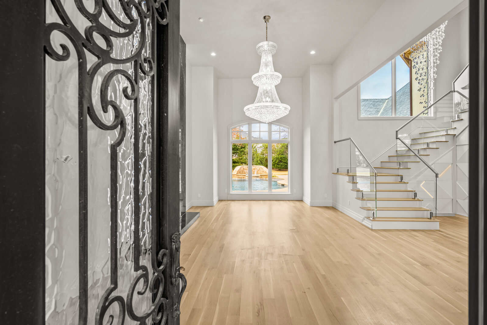 Open foyer with a massive multi-tiered crystal chandelier, modern open staircase with glass railings and wood treads, view through to the pool through a tall arched window