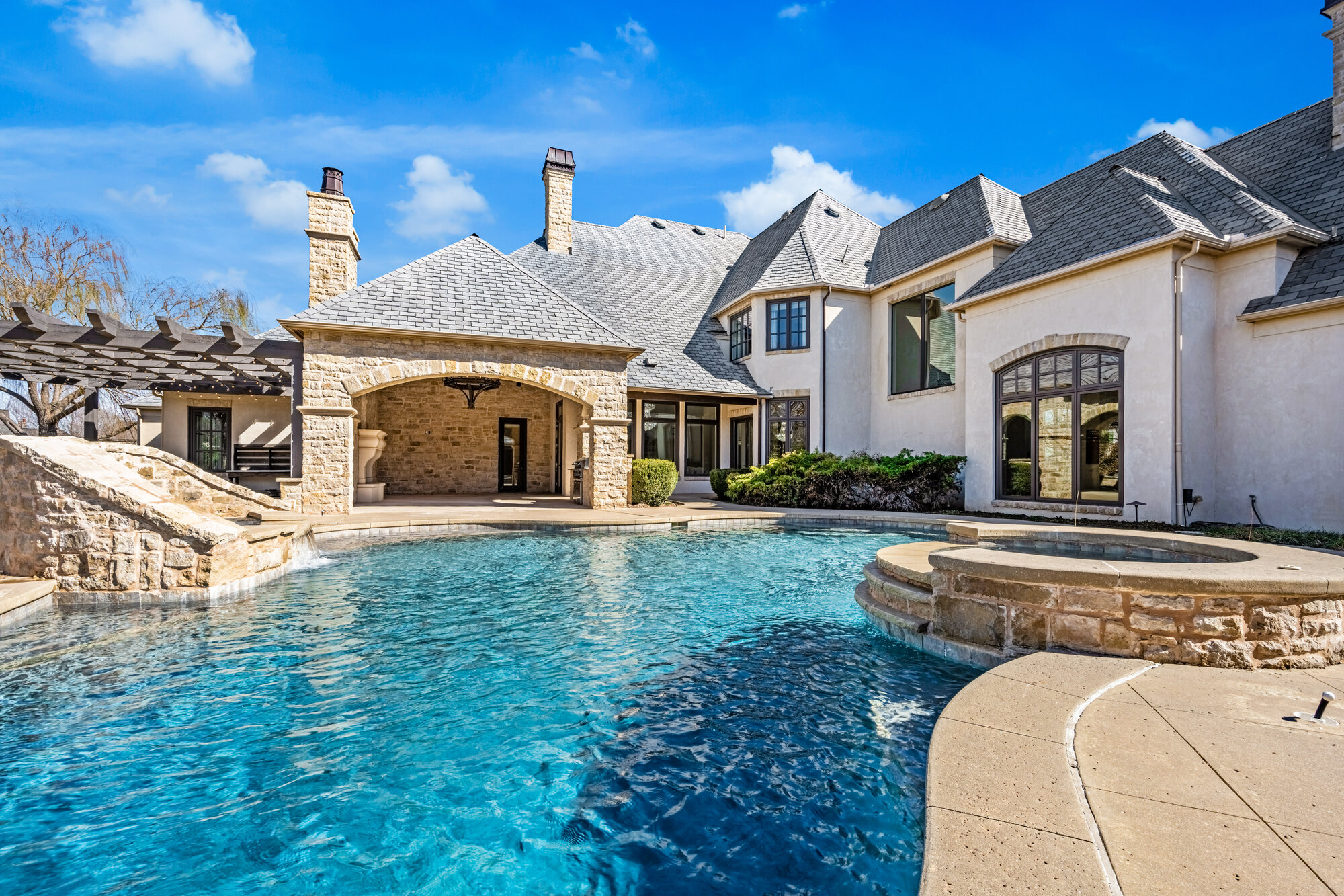 Rear of the estate with a custom shaped pool and attached spa, stone waterfall feature, stone pergola on the left, and a covered stone pavilion attached to the home