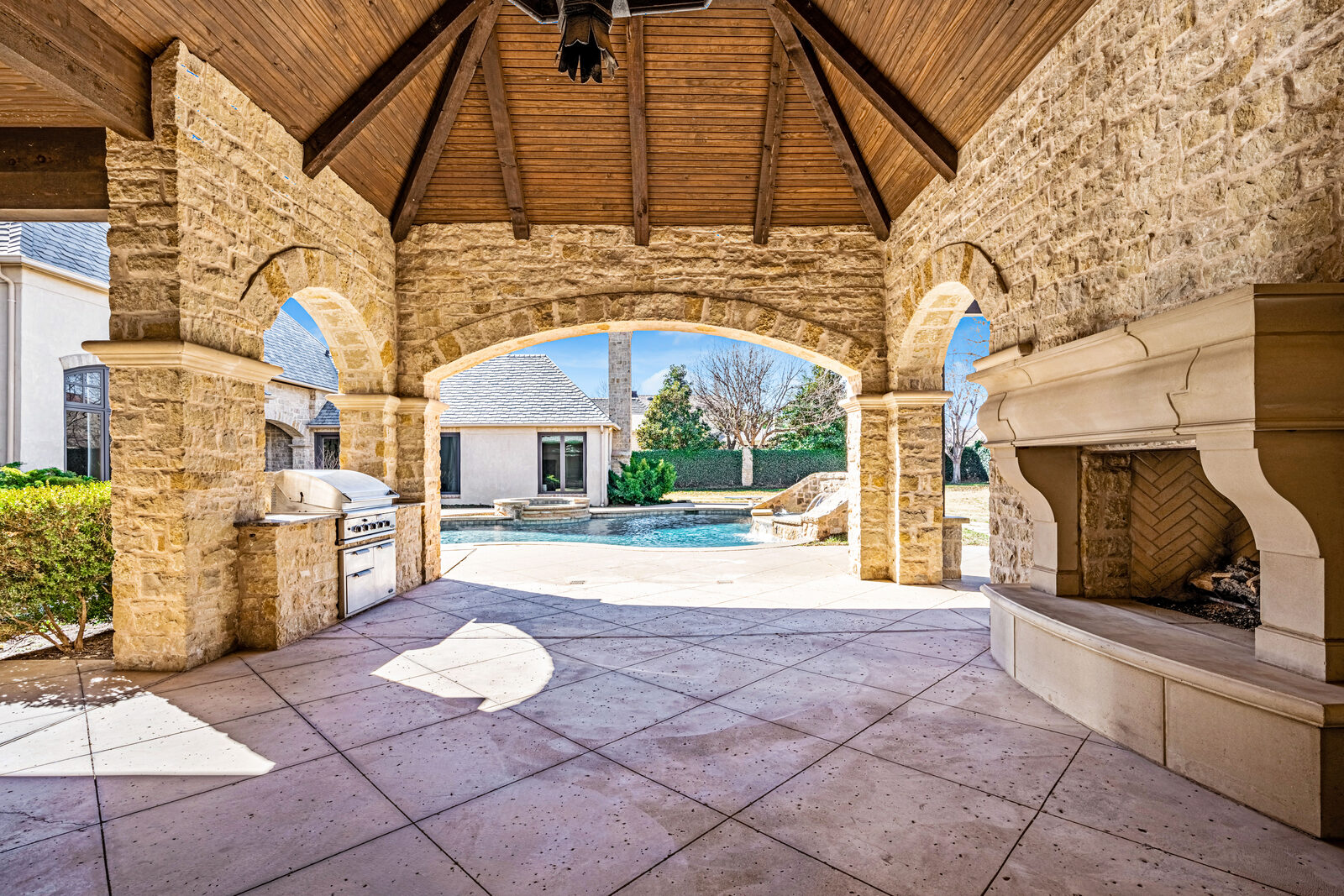 Covered outdoor stone pavilion with vaulted timber ceiling, stone arches framing the pool view, built-in stainless gas grill, and a massive carved limestone outdoor fireplace
