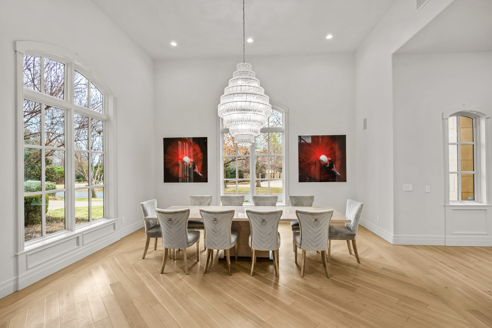 Formal dining room with tiered crystal chandelier, long modern table seating ten, tufted velvet chairs, arched window, and herringbone-pattern oak floors