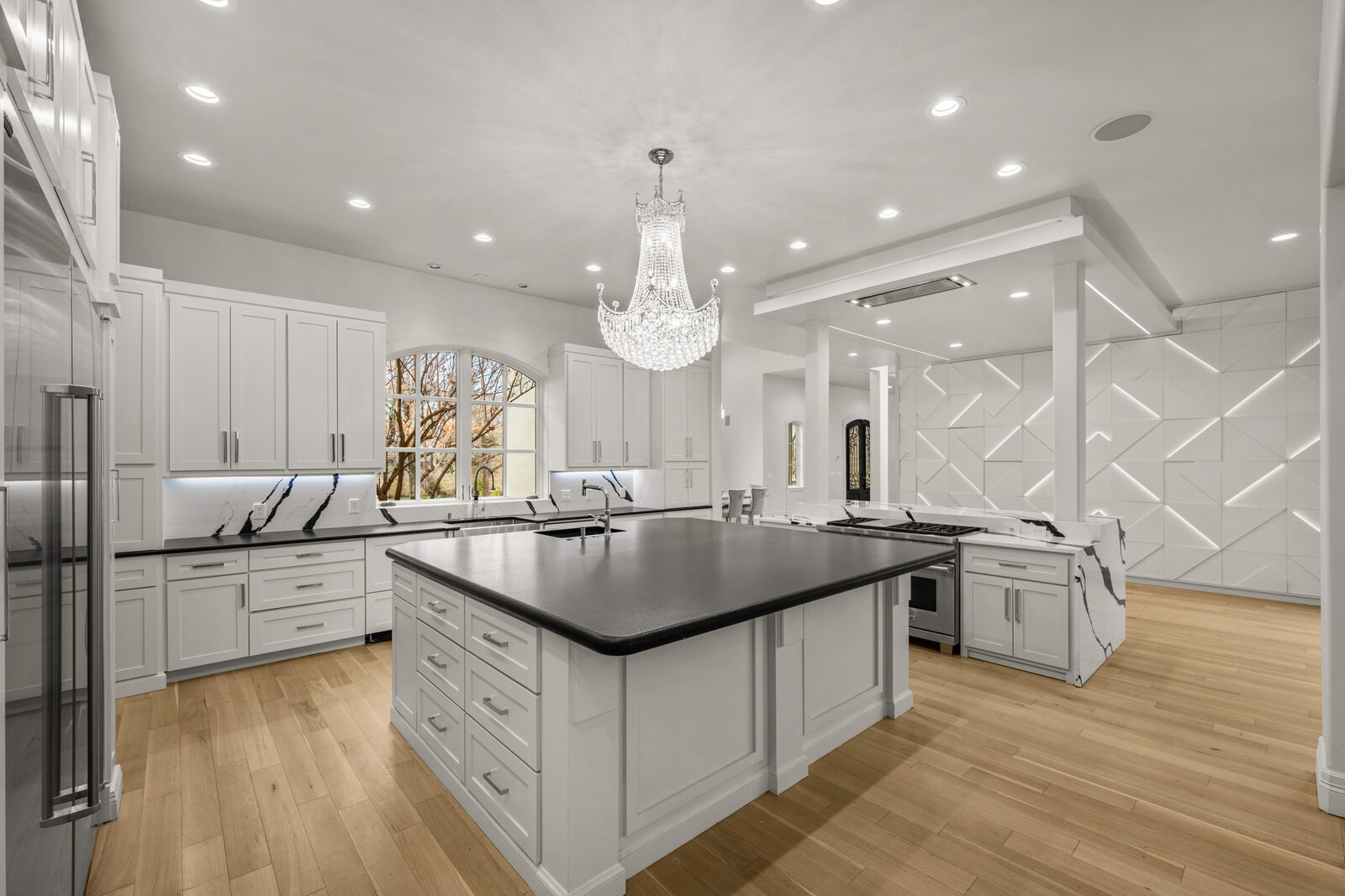 Chef's kitchen with dual islands — central island with black soapstone, perimeter with white Calacatta marble counters, professional stainless cooktop with downdraft vent, 3D textured accent wall behind the cooking station, crystal chandelier overhead