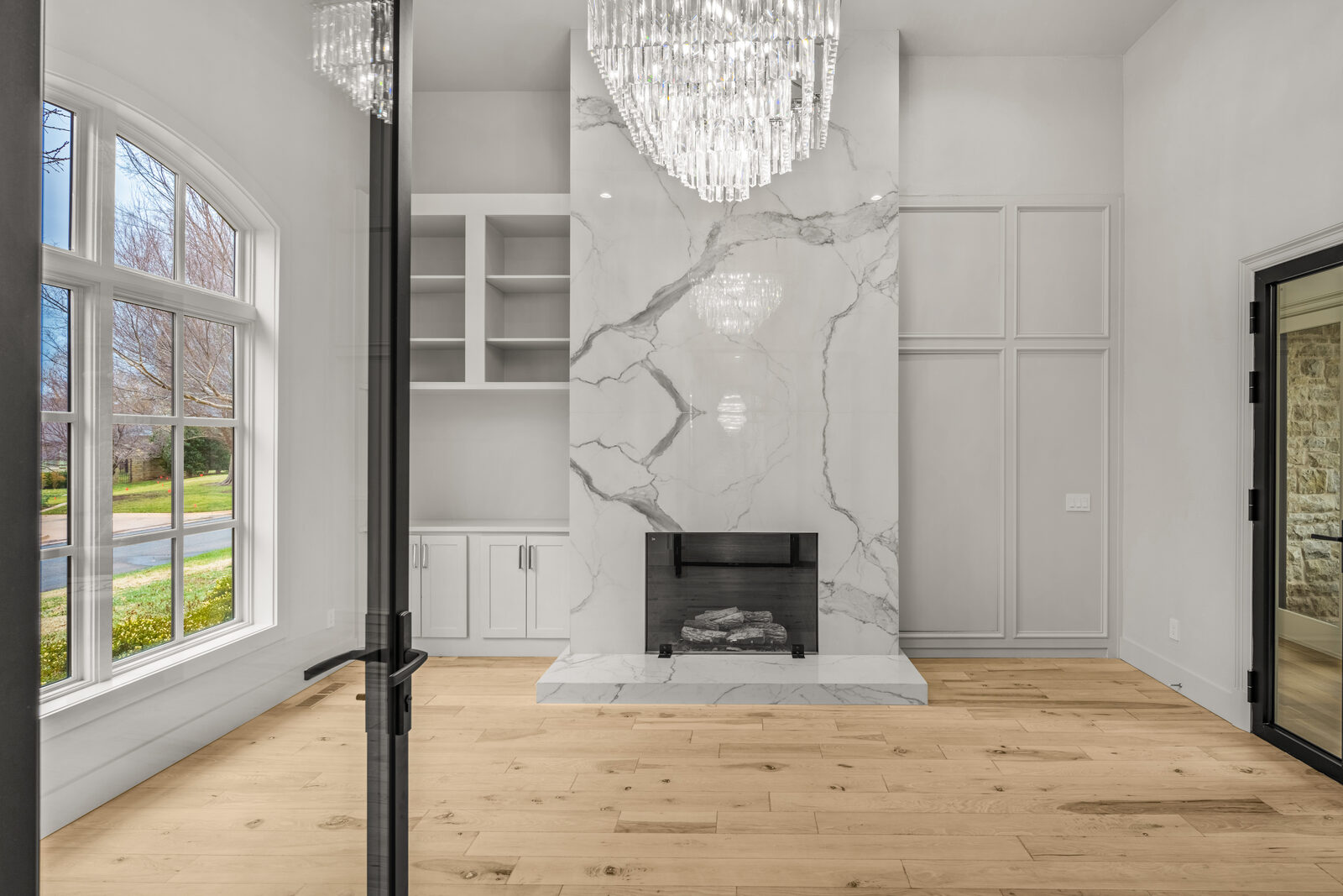 Formal sitting room with a floor-to-ceiling white Calacatta marble fireplace, built-in white cabinetry, cascade crystal chandelier, tall arched window with views of the front grounds