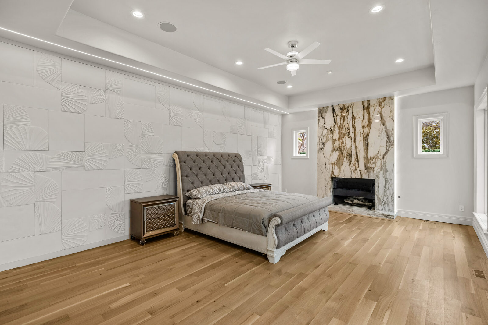 Primary bedroom with a 3D textured palm-fan accent wall, cream-and-gold marble fireplace, tufted gray velvet sleigh bed, tray ceiling with integrated LED accent lighting, light oak floors