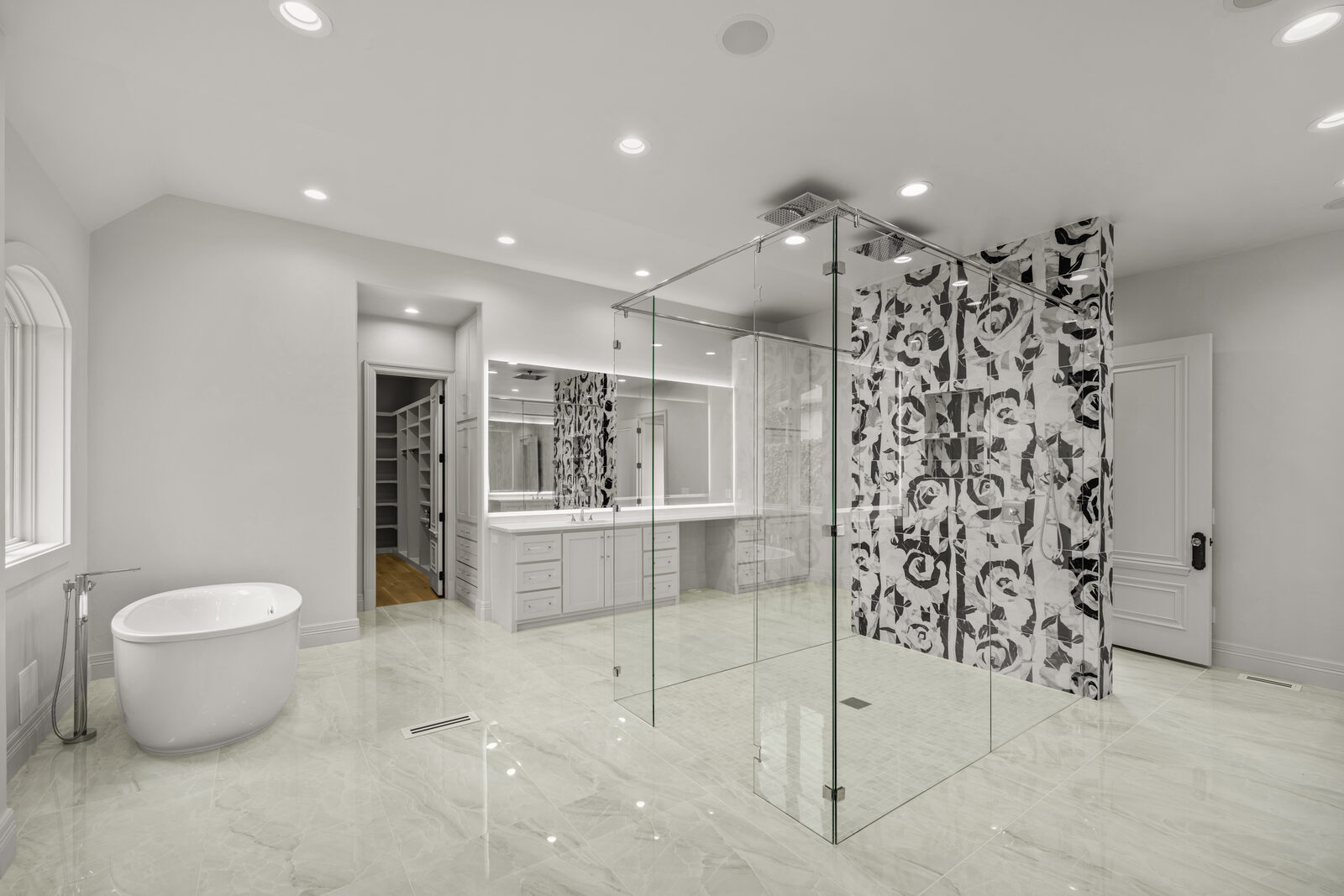 Primary bath with a centered glass-enclosed shower wrapped in bold black-and-white rose-pattern tile, freestanding modern soaking tub on the left, polished marble-look floor tile, LED-backlit mirror over the vanity
