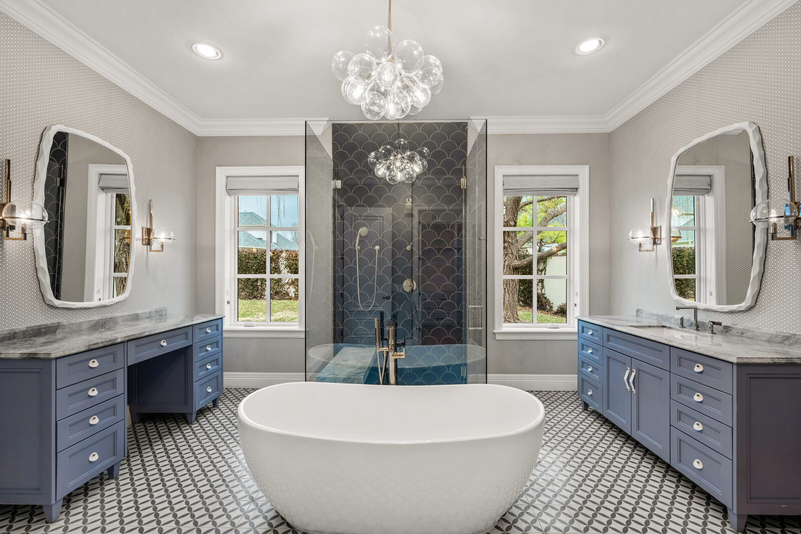 Designer guest bath with navy blue cabinetry, navy fish-scale shower tile, patterned black-and-white geometric floor, freestanding textured soaking tub, glass bubble chandelier, and brass sconces