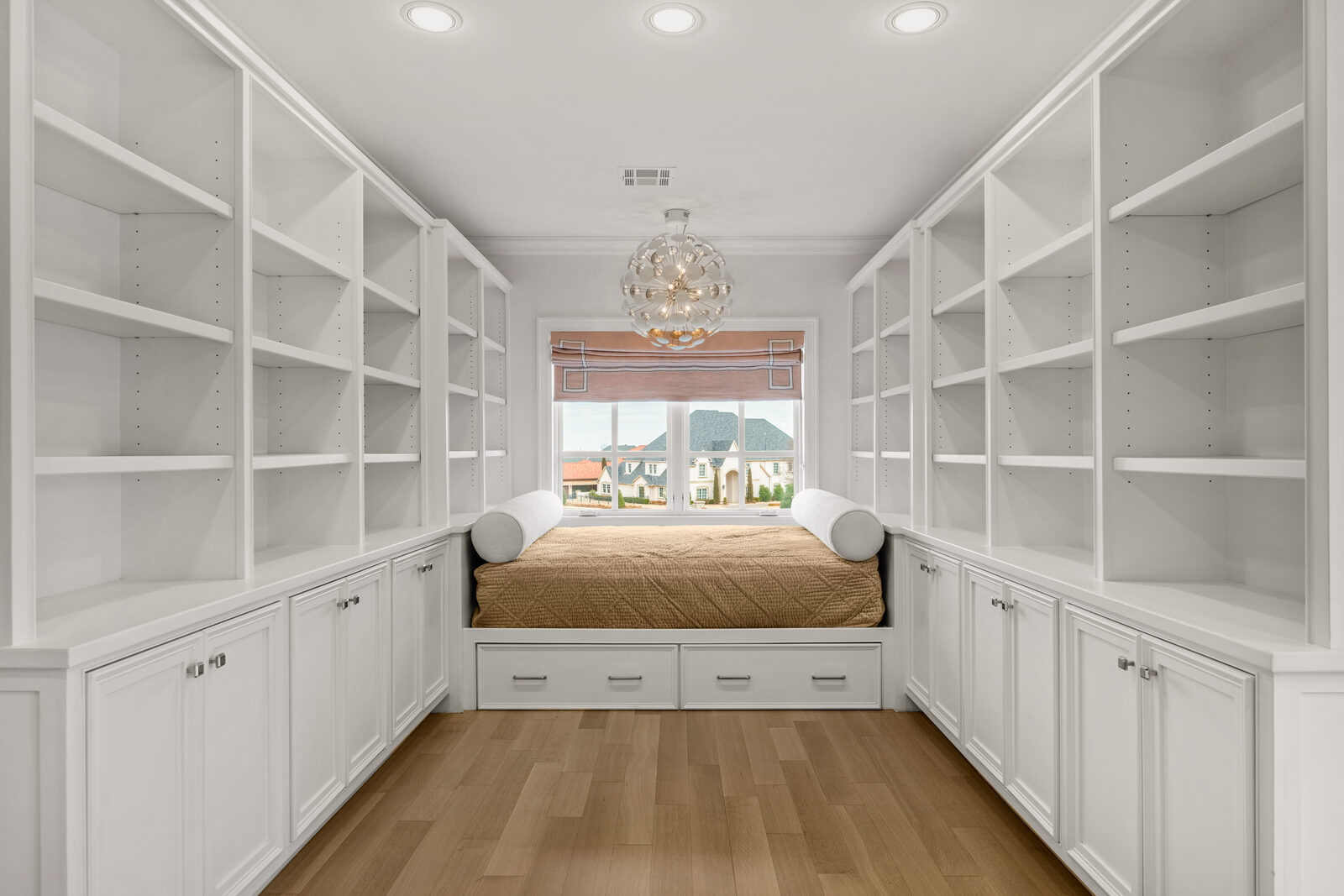 Symmetrical library or reading nook with floor-to-ceiling built-in white shelving on both sides, built-in window day bed, glass orb chandelier, and views to other estates in the neighborhood